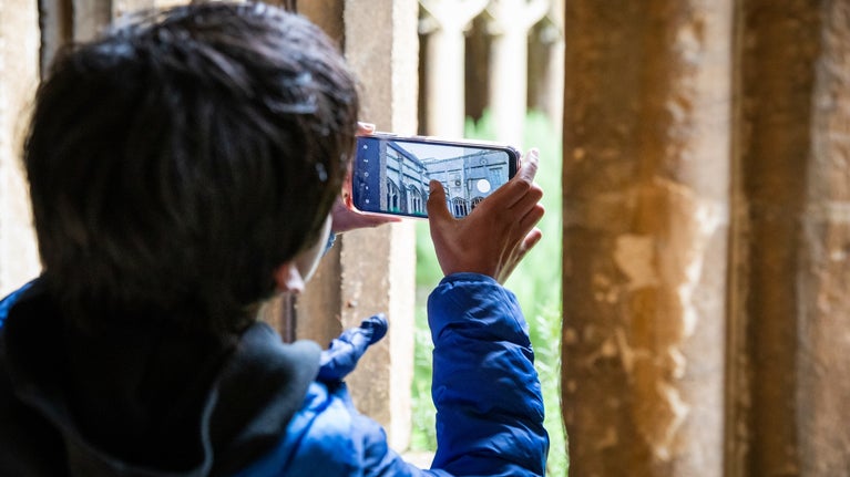 A child gets ready to snap a photo on their phone through the window of the Cloister at Lacock, they use their thumb to focus the image which captures the stone walkway across the Cloister Garth.